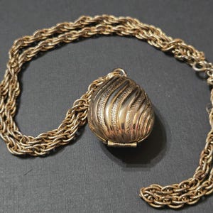 Könnte beinhalten: Eine goldfarbene Metallkette mit einer dicken, gedrehten Kordelkette und einem zentralen, tropfenförmigen Medaillonanhänger. Das Medaillon hat ein strukturiertes, wirbelndes Rippenmuster auf der Oberfläche und ein kleines Scharnier am unteren Rand.