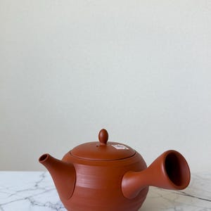 Puede incluir: Una tetera japonesa kyusu de color terracota con un asa lateral y una tapa a juego. La tetera presenta un acabado liso y mate con un cuerpo redondeado. Se puede observar una pequeña pegatina cuadrada sobre la tapa de la tetera.