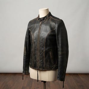 May include: A dark brown leather jacket with a distressed finish, featuring a central metal zipper, a mandarin collar with a snap button closure, long sleeves with zippered cuffs, and two vertical zippered pockets on the front torso area.