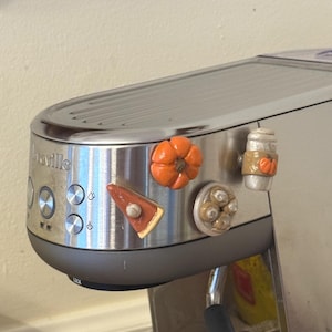 Peut inclure: Une machine à expresso Breville argentée avec quatre petits aimants décoratifs sur le thème de l'automne fixés sur le panneau avant. Les aimants représentent une citrouille orange, une part de tarte, un biscuit rond et une tasse à café blanche.