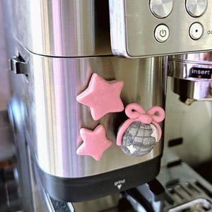 May include: Two smooth, light pink star-shaped magnets and one round, silver glittery disco ball magnet topped with a pink bow, all attached to the brushed stainless steel surface of a coffee machine with visible power and grind buttons.