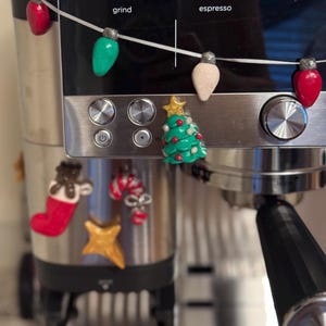 May include: A Ninja espresso machine decorated with holiday-themed magnets including a string of colorful light bulbs, a green Christmas tree with a gold star, a red stocking with a brown bear, a candy cane, and a gold star.
