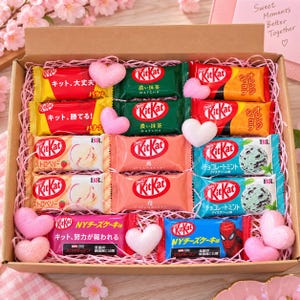 May include: A cardboard gift box filled with assorted KitKat chocolate bars in various flavors and packaging designs, including banana, matcha, orange, strawberry, chocolate mint, and New York cheesecake, accented with small pink foam hearts.