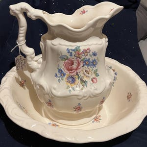 May include: A cream-colored ceramic wash basin and matching pitcher set featuring embossed scalloped edges and a floral pattern of pink, yellow, and blue flowers. The pitcher has a decorative handle and a tag with handwritten text attached.