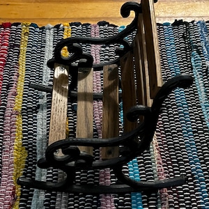 Op de afbeelding: Een miniatuur schommelstoel met een zwart metalen frame met sierlijke krulvormige armleuningen en rugsteunen. De zitting en rugleuning zijn opgebouwd uit horizontale houten latten die met zichtbare metalen schroeven aan het frame zijn bevestigd.