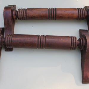 May include: Two antique wooden towel bars. Each bar is a dark brown color and features a cylindrical center with carved ridges and decorative end pieces. The bars are designed to be mounted on a wall.