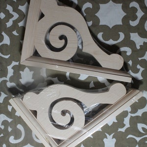 May include: Two unfinished wooden corner brackets with a decorative scroll design. The brackets are wrapped in plastic and are ready to be painted or stained.
