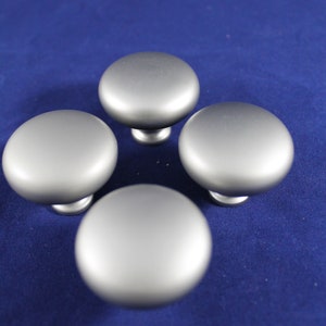 May include: Four silver cabinet knobs with a smooth, rounded surface. The knobs are arranged in a diamond shape on a blue background.