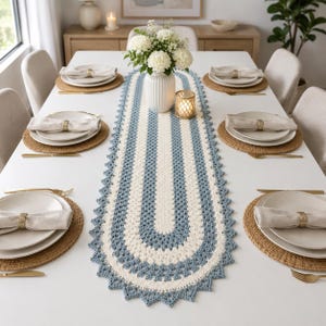 May include: An oval crochet table runner featuring alternating bands of cream and dusty blue yarn with a scalloped edge. The runner is centered on a white dining table set with four place settings, each including a round woven placemat, white plates, and a linen napkin held by a gold ring.