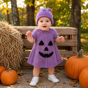 Può includere: Un vestito in maglia viola con cappellino abbinato, caratterizzato da un disegno nero a forma di faccia di zucca sul davanti. Il vestito ha maniche corte e uno scollo a coste, mentre il cappellino presenta un piccolo dettaglio a stelo verde.