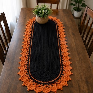 May include: An oval-shaped crochet table runner featuring a solid black centre with a textured ribbed pattern, surrounded by a wide, intricate orange border with a scalloped edge design. The runner is laid flat on a wooden surface.