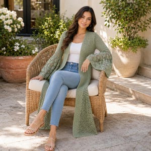 Könnte beinhalten: Eine salbeigrüne, offene Strickjacke mit langen Glockenärmeln, aufwendigen Häkeldetails, einem schlichten Strickoberteil und langem Saum, kombiniert mit einem weißen Tanktop, einer hellblauen Jeans und goldenen Riemchensandalen.