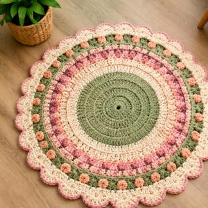 Può includere: Un tappeto rotondo all'uncinetto con cerchi concentrici nei colori verde salvia, crema e varie tonalità di rosa. Il design presenta punti a nocciolina testurizzati e un bordo smerlato rifinito con piccoli pon-pon rosa chiaro.