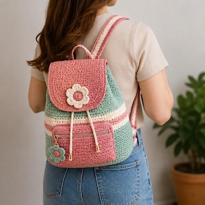 Puede incluir: Una mochila de ganchillo con una solapa rosa que presenta una aplicación de flor blanca, un cuerpo a rayas verdes y blancas, y un bolsillo frontal con cremallera. Incluye cordones blancos con puntas metálicas doradas y un pequeño dije de flor.