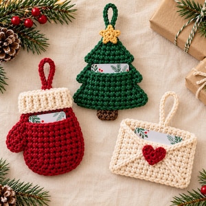 Puede incluir: Tres adornos de ganchillo con lazos para colgar, diseñados como una manopla roja con puño crema, un árbol de Navidad verde con una estrella amarilla y un sobre crema con un corazón rojo, cada uno con una pequeña tarjeta con estampado botánico.