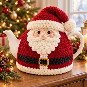 Puede incluir: Una tetera de cerámica blanca cubierta por una funda de ganchillo con forma de Papá Noel. La funda presenta un gorro rojo con un pompón blanco, una barba blanca, un cinturón negro con hebilla amarilla y un borde blanco en la base.