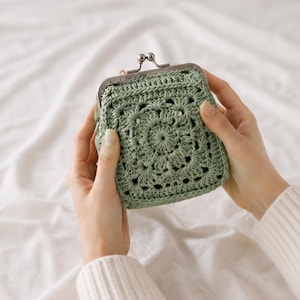 Puede incluir: Un monedero de ganchillo en color verde salvia con un patrón de mandala cuadrado y un cierre de boquilla metálico en tono plateado. La bolsa está confeccionada con puntadas de hilo intrincadas, creando una superficie texturizada y circular.