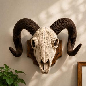 May include: A crochet ram skull wall mount featuring a cream-colored skull and large, textured dark brown curved horns. The skull is securely attached to a dark wooden plaque with a scalloped edge, designed for display as a decorative wall piece.