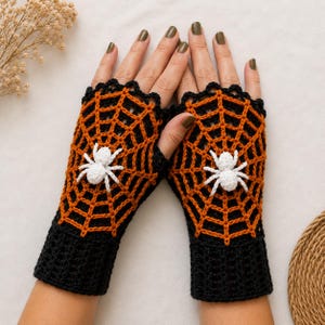 Puede incluir: Un par de guantes sin dedos con un patrón de telaraña de ganchillo en negro y naranja. Cada guante está adornado con una pequeña araña blanca de ganchillo en tres dimensiones centrada en el diseño de la red, con un puño acanalado negro.