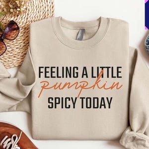 May include: A beige crewneck sweatshirt featuring the black text "FEELING A LITTLE" and "SPICY TODAY" with the word "pumpkin" written in a cursive orange font in the center. The garment has long sleeves and a ribbed collar, cuffs, and hem.