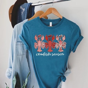 May include: A teal short-sleeved t-shirt featuring a graphic of three stylized red crawfish with patterned details. Below the crawfish, the text crawfish season is printed in a white cursive font. The shirt is displayed on a wooden hanger.