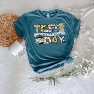 May include: A teal short-sleeve t-shirt featuring a graphic print with the text "TEST DAY" and "MAY THE SKILLS BE WITH YOU". The letters contain illustrations of various space-themed characters, robots, and a spaceship on a dark blue background.