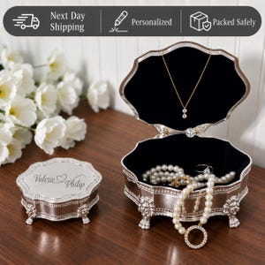 May include: A silver-toned metal jewelry box with a scalloped edge and ornate feet. The lid is engraved with the names Valerie and Philip separated by a heart. The open box features a black velvet interior containing a pearl necklace and a pendant.