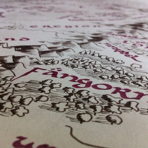 May include: Close-up of a map with the word "Fangorn" in a deep red color. The map is hand-drawn with black ink, depicting a landscape with mountains, trees, and text in a stylized font. The paper has a slightly textured, off-white appearance.