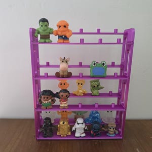 May include: A purple plastic display stand with four horizontal shelves featuring small vertical pegs. The shelves hold a collection of various small character figurines, including superheroes, animals, and science fiction figures in diverse colors.