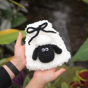 May include: A handmade white crochet bag with a black sheep face and a black bow closure. The sheep has a black face, ears, and a white fluffy body. The bag is held in a person's hands.