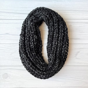 Könnte beinhalten: Ein dunkelgrauer und weiß gesprenkelter Loop-Schal. Der gestrickte Schal ist kreisförmig und wirkt weich und warm. Der Schal ist aus einem dicken Garn gefertigt und wird auf einer weißen Holzoberfläche präsentiert.