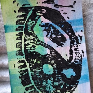 May include: A rectangular print featuring a black ink block-printed yin and yang symbol centered over a background of blended watercolor washes in shades of light green, blue, and purple, creating a textured and layered visual effect.