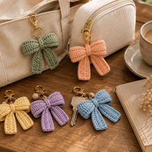May include: Five crochet bow keychains in sage green, peach, pale yellow, lavender, and light blue. Each bow features a gold-toned metal lobster clasp, a small wooden bead, and a textured yarn construction. One blue bow is attached to a metal key.