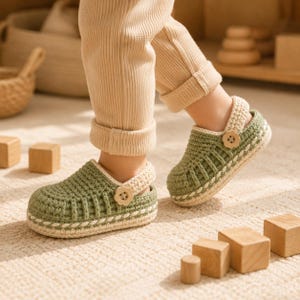 May include: A pair of sage green crochet clogs with a cream-colored heel strap and a small flower-shaped wooden button on the side. The shoes feature a textured knit pattern, a thick cream-colored sole, and a decorative braided trim along the base.