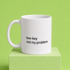 May include: A white ceramic mug with a handle on the left side. The text "low-key not my problem." is printed in black, sans-serif font on the front of the mug. The mug has a smooth, cylindrical body and a simple, functional design.