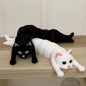 May include: Two articulated cat figurines, one black and one white, featuring segmented bodies, pink-painted ears and noses, and large eyes. The black cat is positioned behind the white cat, both in a prone, outstretched pose with front paws extended.