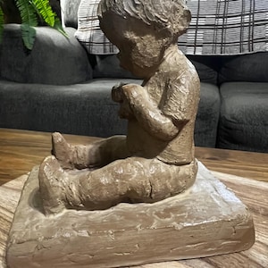 Può includere: Una scultura in argilla marrone e testurizzata di un bambino piccolo seduto su una base quadrata. La figura è raffigurata in posizione seduta con le gambe distese in avanti, indossando una maglietta a maniche corte e dei pantaloni.
