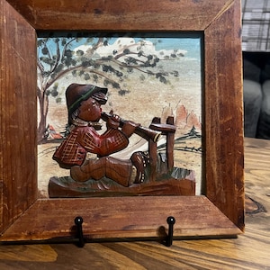 May include: A square wooden wall plaque featuring a relief carving of a person in a hat and plaid jacket playing a flute. The figure sits near a wooden fence in a painted landscape with a tree. The entire scene is enclosed in a brown wooden frame.