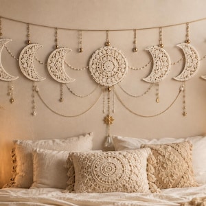 May include: A cream-colored macrame wall hanging featuring a central circular mandala design flanked by six crescent moon shapes. The piece is adorned with hanging gold-toned chains, small beads, and star charms, suspended from a horizontal cord.