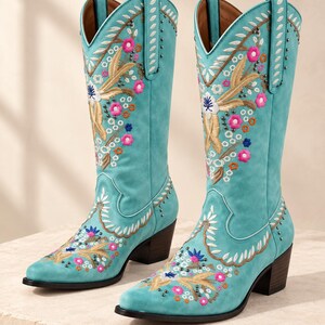 May include: A pair of turquoise mid-calf boots featuring intricate floral embroidery in shades of pink, white, and tan, accented with small metallic studs. The boots have a pointed toe, a stacked block heel, and pull tabs at the top openings.