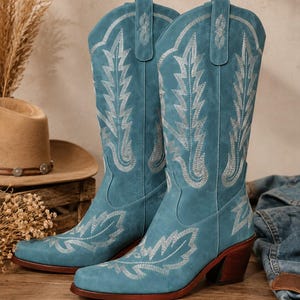 May include: A pair of teal blue mid-calf boots featuring intricate silver-toned embroidery in leaf-like patterns. The boots have a pointed toe, a stacked wooden heel, and pull tabs at the top of the shaft. The exterior has a matte finish.