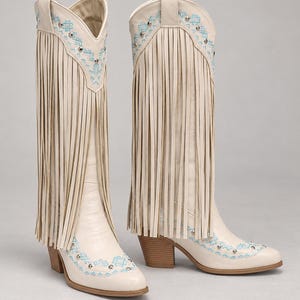 May include: A pair of off-white knee-high boots featuring long fringe detailing along the shaft, light blue geometric embroidery, and silver-toned metal studs. The boots have a pointed toe, a stacked block heel, and a pull-on style design.