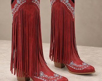 May include: A pair of red knee-high boots featuring long fringe detailing along the sides. The boots are decorated with intricate white and blue geometric embroidery patterns and small silver studs around the collar and the pointed toe area.