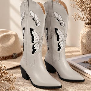 May include: A pair of light gray knee-high boots featuring a pointed toe, a stacked block heel, and a side zipper closure. The boots are decorated with embroidered white and black butterfly wing patterns along the shaft and pull tabs.