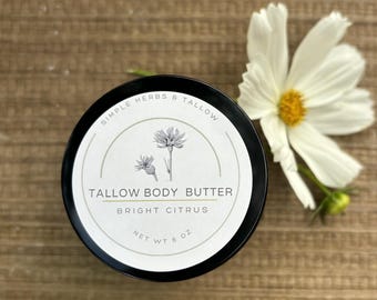 May include: A black circular jar of tallow body butter with a white label featuring a botanical illustration of a flower, the text Simple Herbs & Tallow, Tallow Body Butter, Bright Citrus, and Net Wt 6 oz, accompanied by a white flower.