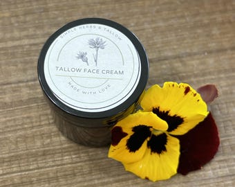 May include: A small black jar of tallow face cream with a circular white label featuring a botanical illustration, the text SIMPLE HERBS & TALLOW, TALLOW FACE CREAM, and MADE WITH LOVE, accompanied by a vibrant yellow and deep maroon pansy flower.