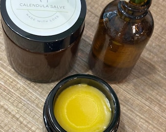 May include: A dark amber glass jar of Calendula Salve with a white label reading Simple Herbs & Tallow, a second open jar revealing a smooth yellow balm, and a small amber glass bottle containing white flowers with green stems and leaves.