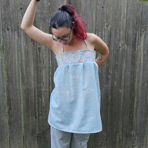 May include: A light blue top with a floral patterned neckline and straps. The top has a light blue body with a subtle floral pattern. The person is wearing gray sweatpants. The background is a wooden fence.