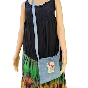 May include: A light blue denim crossbody bag with a rectangular patch featuring a red flower design. The bag has a long strap and is worn over a black dress with a tie-dye pattern in green, orange, and yellow.