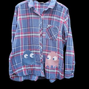 May include: Blue plaid button-down shirt with pink and white accents. Features two pixelated ghost patches, one blue and one pink. The shirt is hanging on a wooden hanger.
